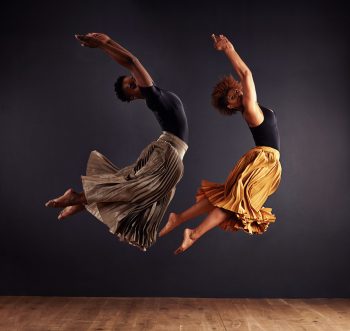 Two contemporary dancers performing a synchronized leap in front of a dark background.